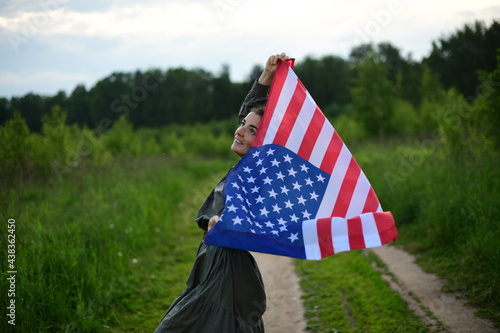 A young, happy American woman with long hair, holding the US national flag fluttering in the wind on her shoulders, is relaxing in the open air, enjoying a warm summer day. July 4