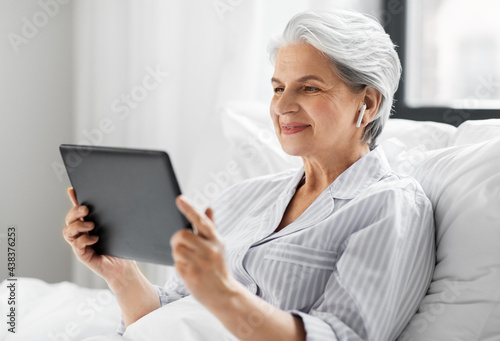 technology, old age and people concept - senior woman with tablet pc computer and wireless earphones in bed at home bedroom