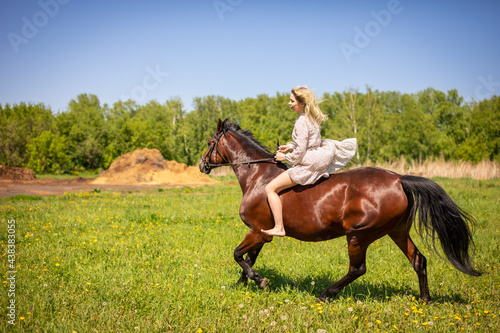 A young rider woman blonde with long hair in a dress riding gallop on brown horse on a field and forest background, Russia