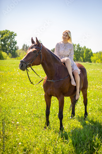 A young rider woman blonde with long hair in a dress posing with brown horse on a field and forest background, Russia © dtatiana