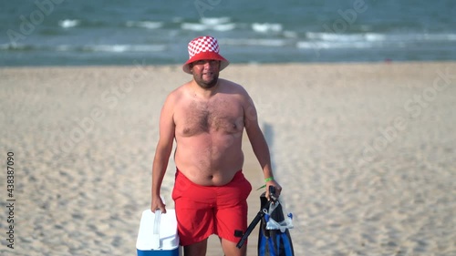 Man in red swimsuit and hat walking on the beach with blue cooler photo