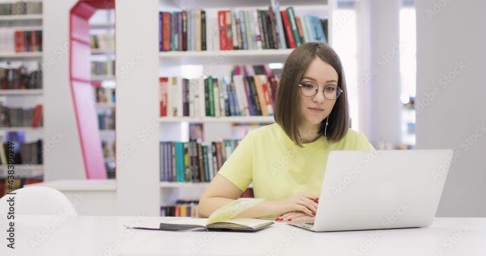 Young woman is sitting at a desk with a laptop, working in the library. She communicates via the Internet. Front view. In the background is a rack with books. Distance learning concept. Young woman is sitting at a desk with a laptop, working in the library. She communicates via the Internet. Front view. In the background is a rack with books. Distance learning concept.