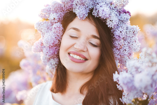 Beautiful happy young woman wear lilac floral wreath over nature background closeup. Attractive girl smiling outdoors. Healthy skin and dental smile. 20s. Relaxation.