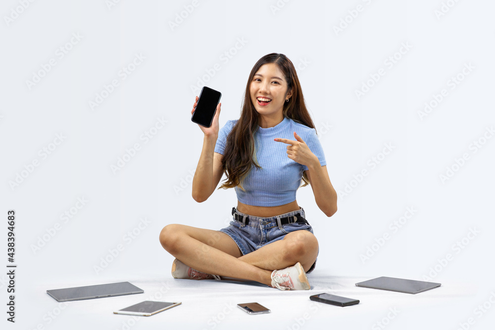 Cheerful Asian woman pointing at smartphone with black screen and sitting on white background with digital device Cheerful Asian woman pointing at smartphone with black screen and sitting on white background with digital device