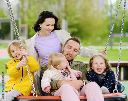 Merry family with three little children having fun on outdoor playground. Young parents rides siblings on swing. Spring/summer/autumn active leisure for family with kids.