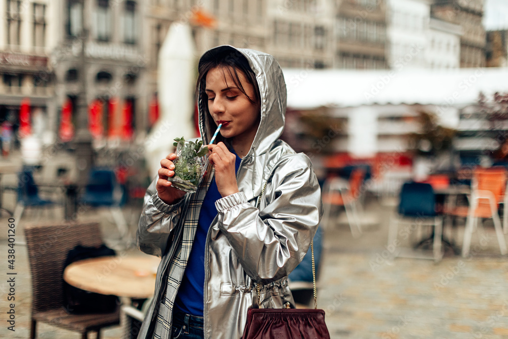 girl drinking mojito with straw in city center. young woman in raincoat having a drink in happy hour.