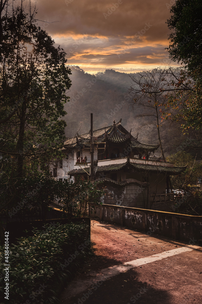 Obraz premium On way to Qingcheng mountain, Sichuan, China. Breathtaking golden hour sunset over mountains and old Asian Chinese style building house near road path. Traditional architecture and spears on roof edge