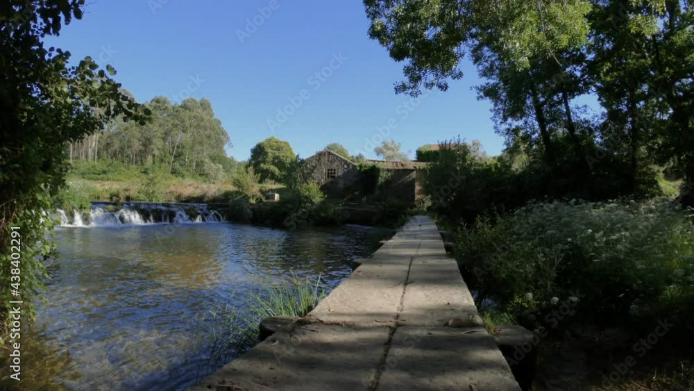 SLOW MOTION SHOT - Old stone footbridge over the Neiva River in Antas, Esposende, Portugal with an old Azenhas, ancient mills, in the background. SLOW MOTION SHOT - Old stone footbridge over the Neiva River in Antas, Esposende, Portugal with an old Azenhas, ancient mills, in the background.
