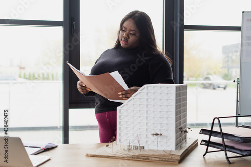 brunette african american plus size engineer holding folder near house model on desk.