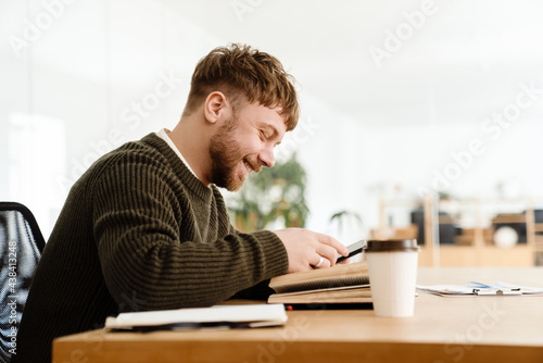 Young ginger man smiling and using mobile phone while reading book © Drobot Dean