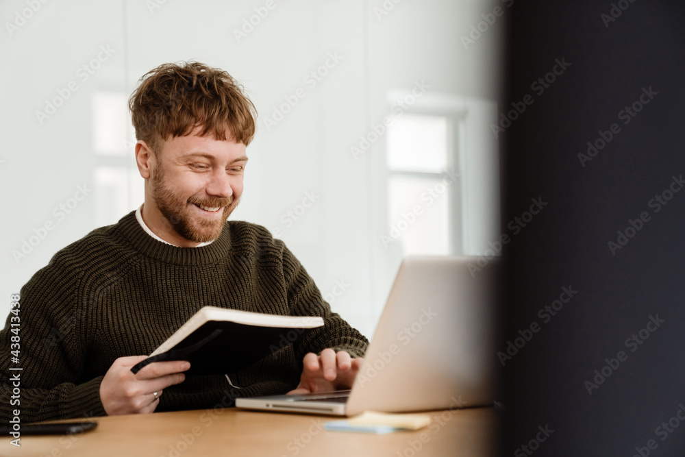 Young ginger man working with laptop while sitting at table in office Young ginger man working with laptop while sitting at table in office