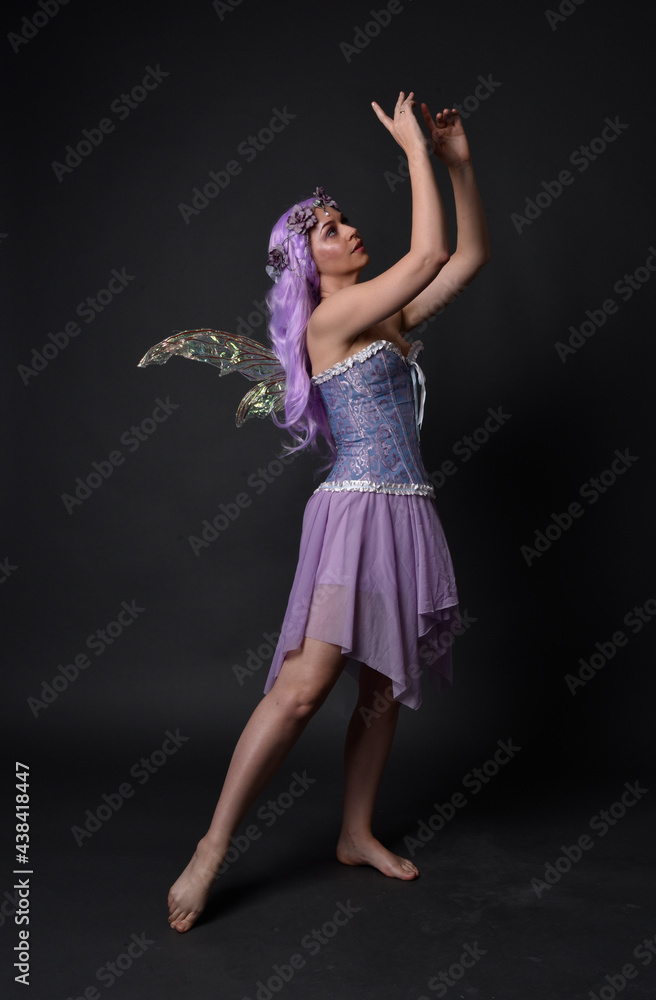 Full length portrait of a purple haired girl wearing fantasy corset dress with fairy wings and flower crown. Standing pose against a dark studio background. Full length portrait of a purple haired girl wearing fantasy corset dress with fairy wings and flower crown. Standing pose against a dark studio background.