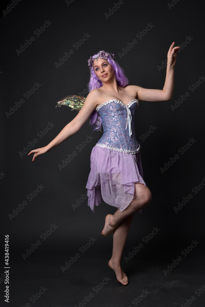 Full length portrait of a purple haired girl wearing fantasy corset dress with fairy wings and flower crown. Standing pose against a dark studio background. Full length portrait of a purple haired girl wearing fantasy corset dress with fairy wings and flower crown. Standing pose against a dark studio background.