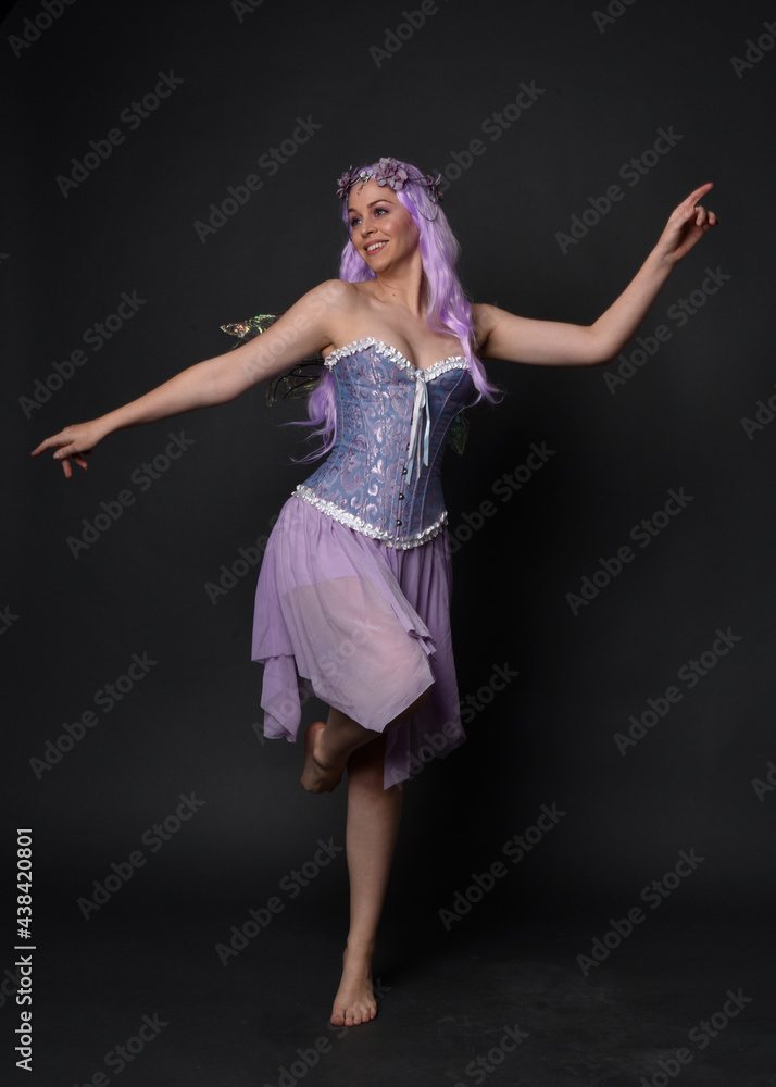 Naklejka premium Full length portrait of a purple haired girl wearing fantasy corset dress with fairy wings and flower crown. Standing pose against a dark studio background.