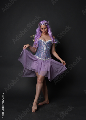 Full length portrait of a purple haired girl wearing fantasy corset dress with fairy wings and flower crown. Standing pose against a dark studio background.