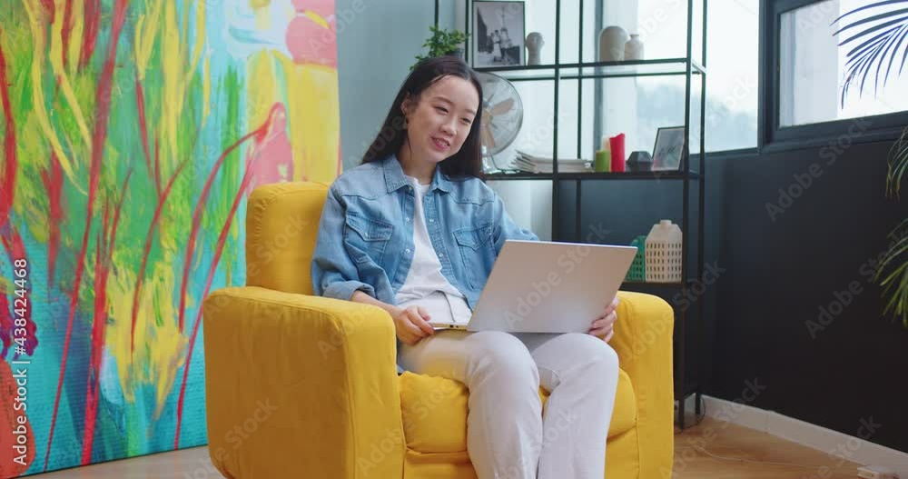 Happy Asian woman videochatting on laptop sitting in living room in chair and waving hand and smiling. Young joyful beautiful female in apartment speaking on online video call on computer Happy Asian woman videochatting on laptop sitting in living room in chair and waving hand and smiling. Young joyful beautiful female in apartment speaking on online video call on computer