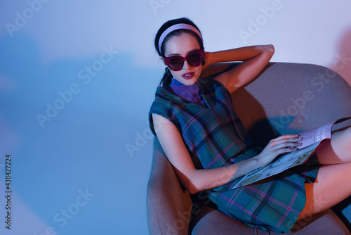 young woman in sunglasses and retro outfit sitting in armchair with magazine on blue.