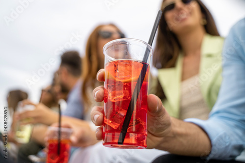 Gathering of best friends having fun talking and drinking cocktails together. Close up of an hand holding an iced spritz drink. Defocused young people on the background.