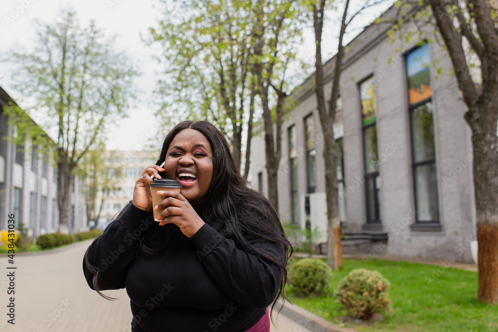 cheerful african american plus size woman holding coffee to go and talking on smartphone while laughing outside. cheerful african american plus size woman holding coffee to go and talking on smartphone while laughing outside.
