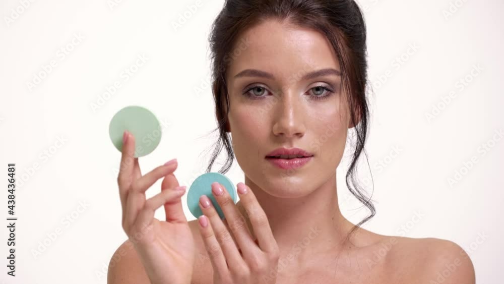 A close-up view of a beautiful halfnaked woman is posing with colorful sponges in her hands standing isolated over white wall in the studio A close-up view of a beautiful halfnaked woman is posing with colorful sponges in her hands standing isolated over white wall in the studio