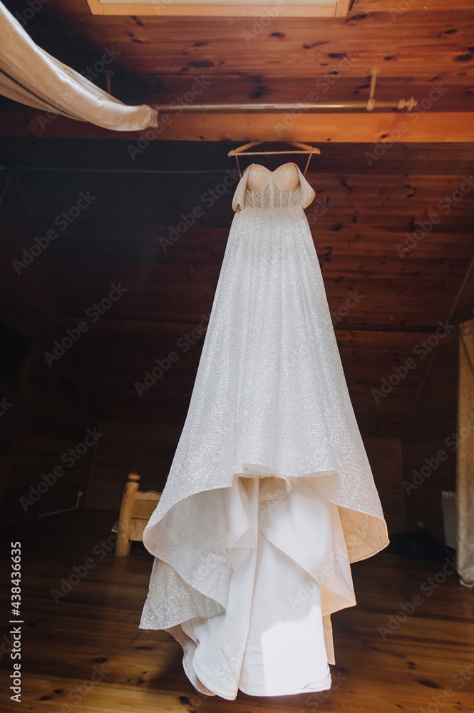 A beautiful, long, white dress of the bride hangs on a hanger against the background of a wooden interior. Photography, concept.