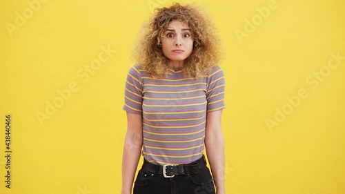 A confused shocked curly blonde woman is standing isolated over yellow wall in the studio photo