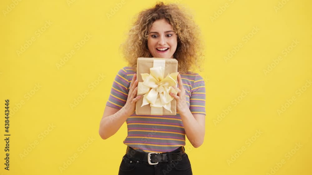 A happy blonde woman is shaking a present box while standing isolated over yellow wall in the studio A happy blonde woman is shaking a present box while standing isolated over yellow wall in the studio