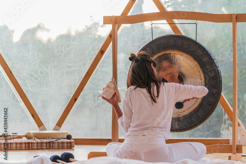 Spiritual woman hitting gong while calm people lying on yoga mats with eyes closed in yoga class
