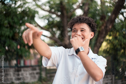 An young mixed race man points to someone, laughing out loud. Comedic situtation or mocking a person. Outdoor background.