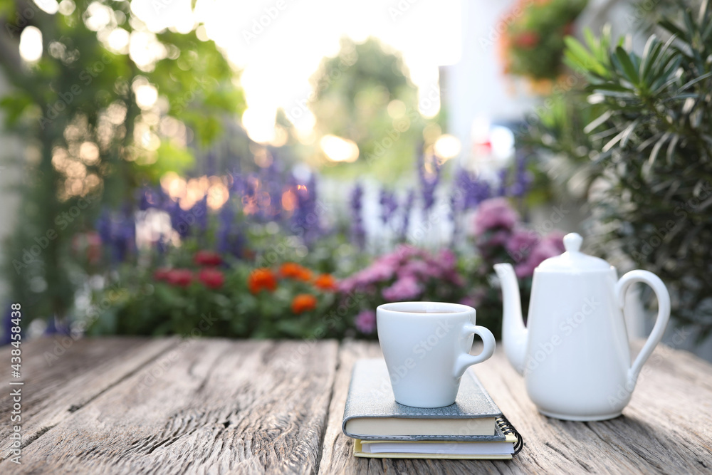 White coffee cup and teapot and notebooks on wooden table with flowers in the back White coffee cup and teapot and notebooks on wooden table with flowers in the back