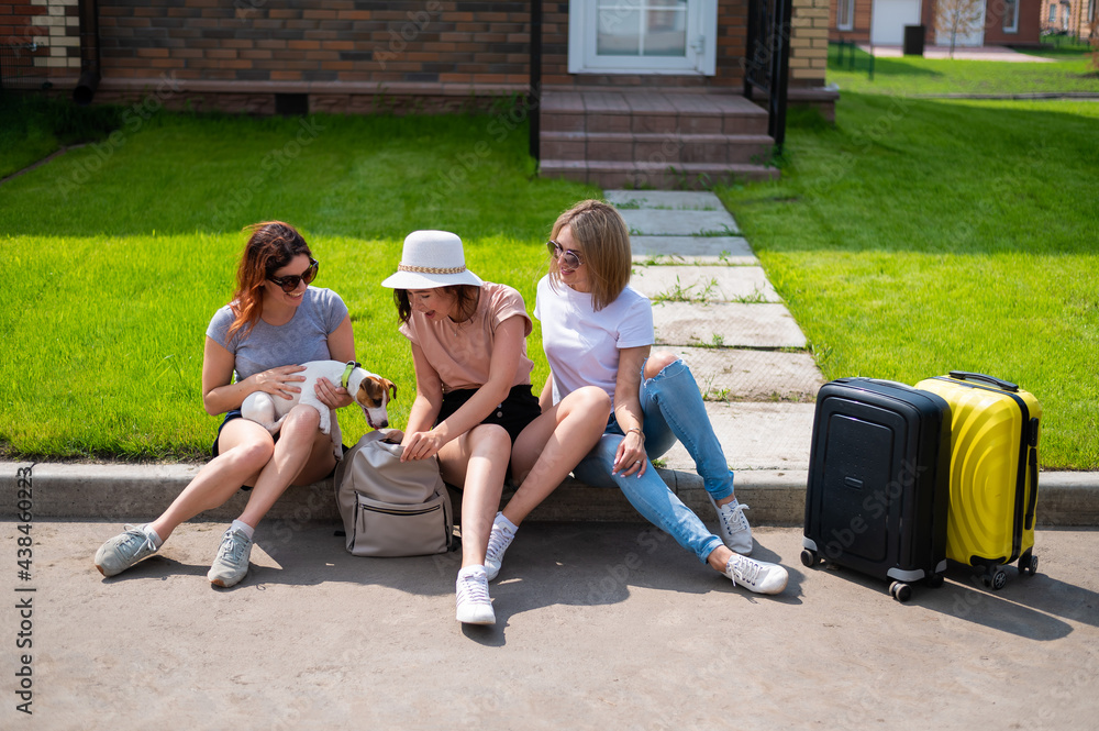 Three Caucasian women and a dog go on a trip. The girls are sitting on the curb with suitcases and waiting for a taxi. Summer vacation concept together with girlfriends Three Caucasian women and a dog go on a trip. The girls are sitting on the curb with suitcases and waiting for a taxi. Summer vacation concept together with girlfriends
