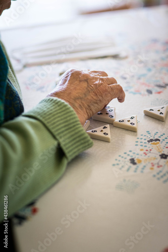 Main d'une femme âgée qui joue à un jeu de société sur une table napée, avec un masque dans le fond