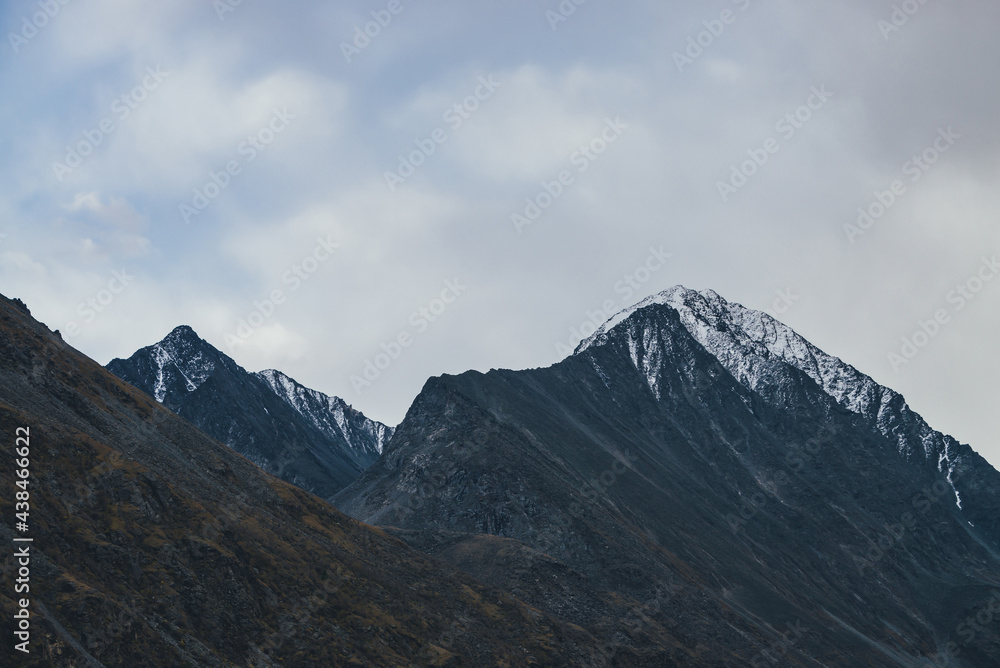 Atmospheric alpine landscape with high mountain silhouette with snow on peaked top under cloudy sky. Dramatic mountain scenery with beautiful snow-covered pointy peak under clouds in overcast weather.