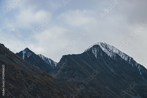 Atmospheric alpine landscape with high mountain silhouette with snow on peaked top under cloudy sky. Dramatic mountain scenery with beautiful snow-covered pointy peak under clouds in overcast weather. © Daniil