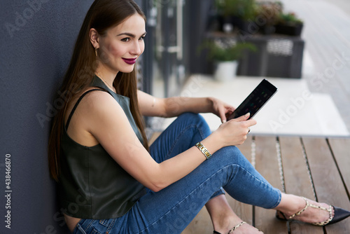 Carefree female influence blogger with modern touch pad gadget resting at urban setting in city and smiling while dreaming about web popularity, pretty hipster girl with digital tablet outdoors