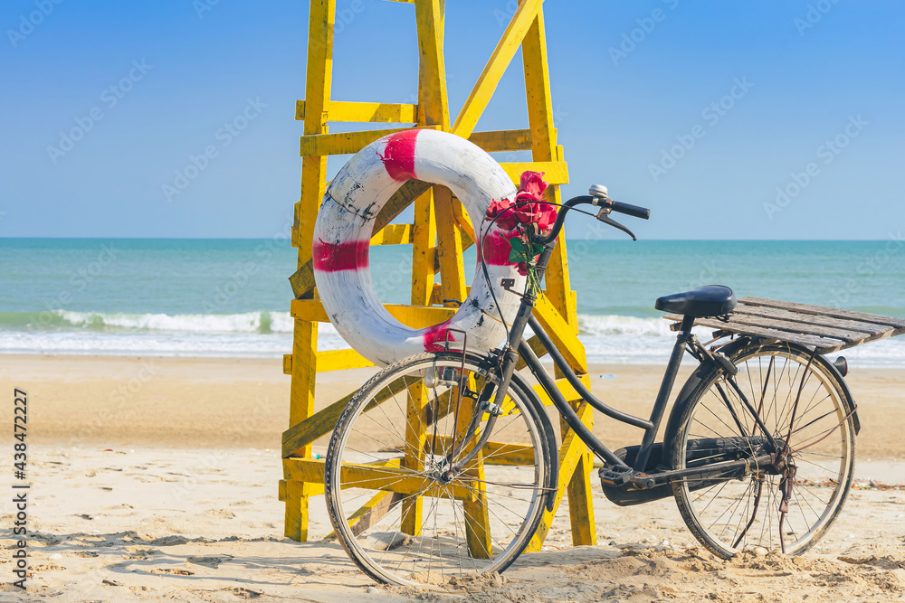 Fototapeta premium Old retro black bicycle with flowers bouquet beside Life ring for life safety on yellow lifeguard stand station or lifeguard chair protecting the safety of tourist on the empty beach in sunshine.