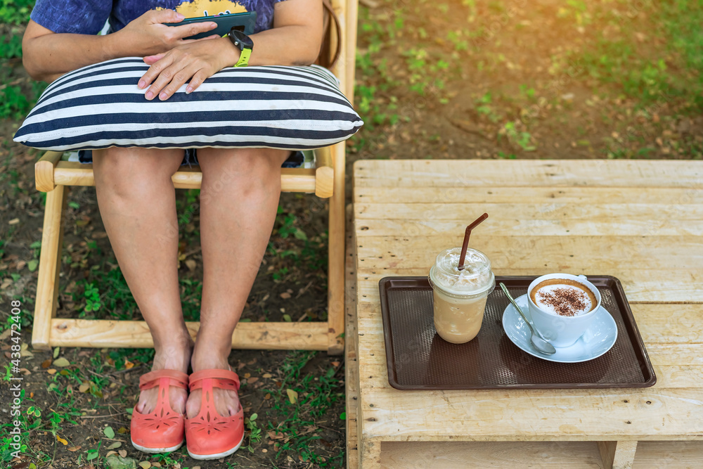 Iced cappuccino coffee and hot cappuccino coffee in brown tray on wooden table for a woman while using her smartphone for a happy relaxing afternoon outdoors in garden. Selective focus on iced coffee.