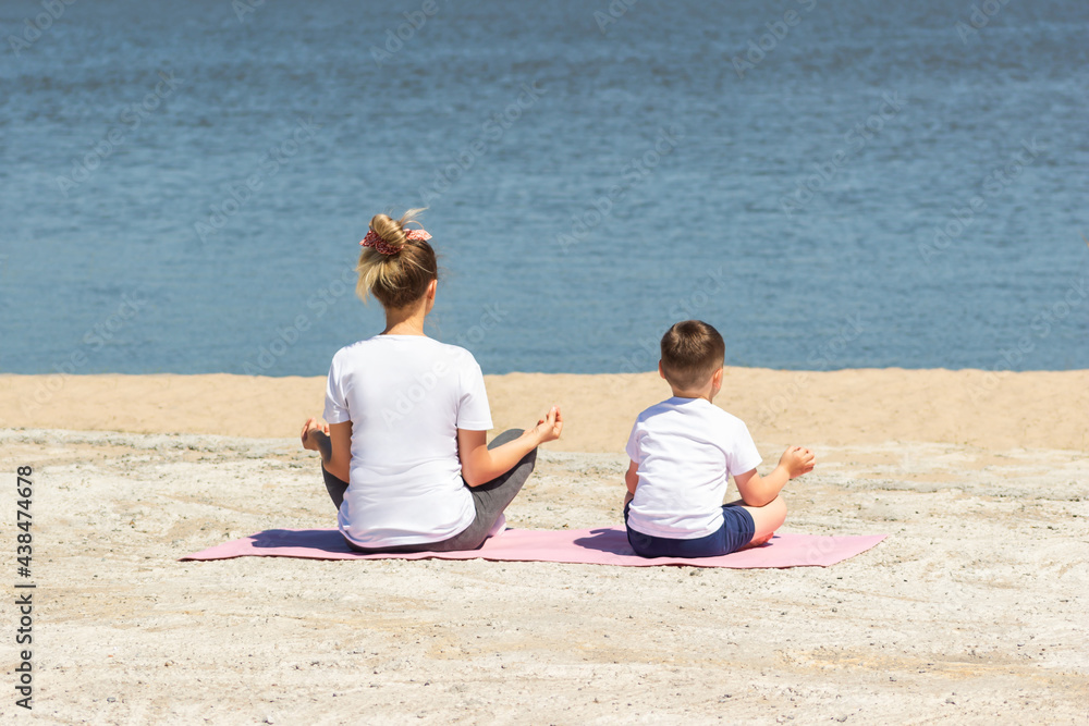 A young woman with her child in white t-shirts meditate holding fingers in yoga sign sitting on a mat by the sea. Mother with her little son in the lotus position. The view from the back. Sunny day
