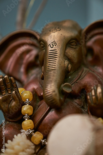 A close up of a statue depicting Oshiro Ganesh with prayer beads and a shamanic drum with teal background. warm tones contrast with cool colors with a lot of detail and interest.