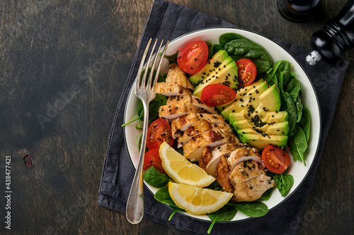 Fresh green salad with grilled chicken fillet, spinach, tomatoes, avocado, lemon and black sesame seeds in white bowl on old wooden dark table background. Nutrition Diet Concept. Top view .