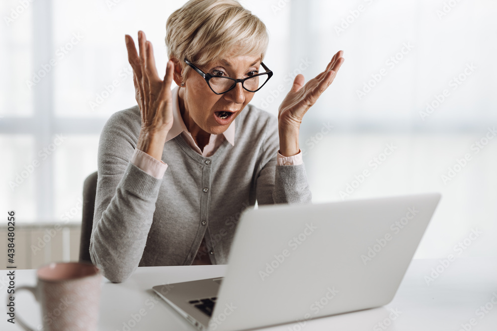 Senior woman reading a shocking e-mail on a laptop in the office Senior woman reading a shocking e-mail on a laptop in the office