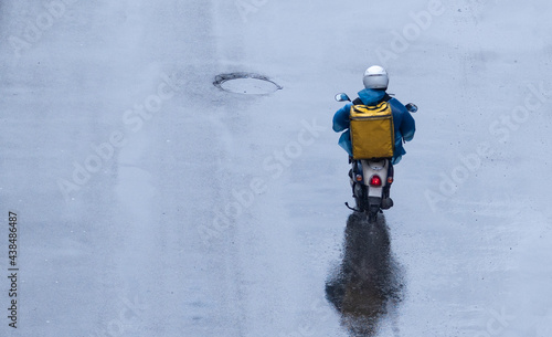 food delivery man in a blue raincoat with a yellow box on his back is eating on wet asphalt