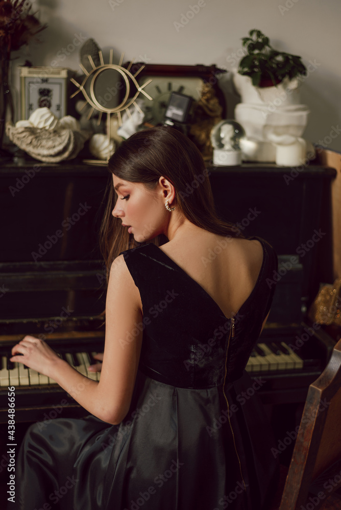 Young attractive lady is posing near piano, wearing black dress. Elegance