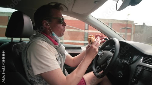 A man takes a bite of a hamburger while driving a car. He holds the steering wheel and a sandwich with his hand, controls the movement of the car. Increased driver energy.
