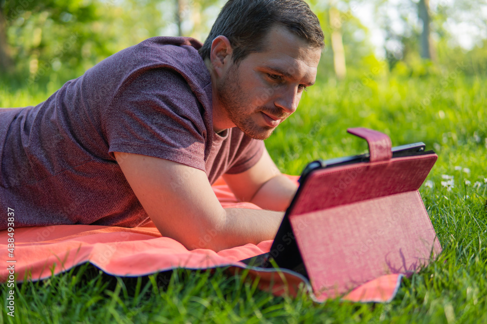 Photo of a young and attractive man sitting in the park on a towel enjoying a sunny day outside in the nature and checking his tablet. Electronic device, technology