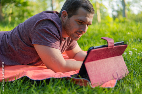 Obraz na płótnie Photo of a young and attractive man sitting in the park on a towel enjoying a sunny day outside in the nature and checking his tablet. Electronic device, technology w salonie