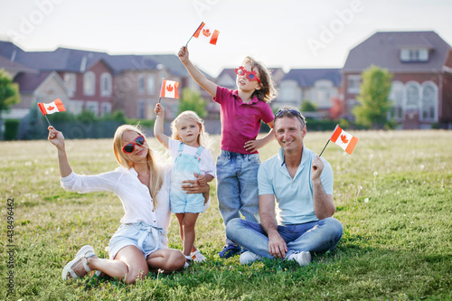 Happy Canada Day. Caucasian family with kids boy and girl sitting on ground grass in park and waving Canadian flags. Parents with kids children celebrating Canada Day on 1st of July outdoor photo