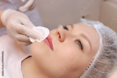 The cosmetologist wipes the remnants of anesthesia from the client's lips with a cotton sponge