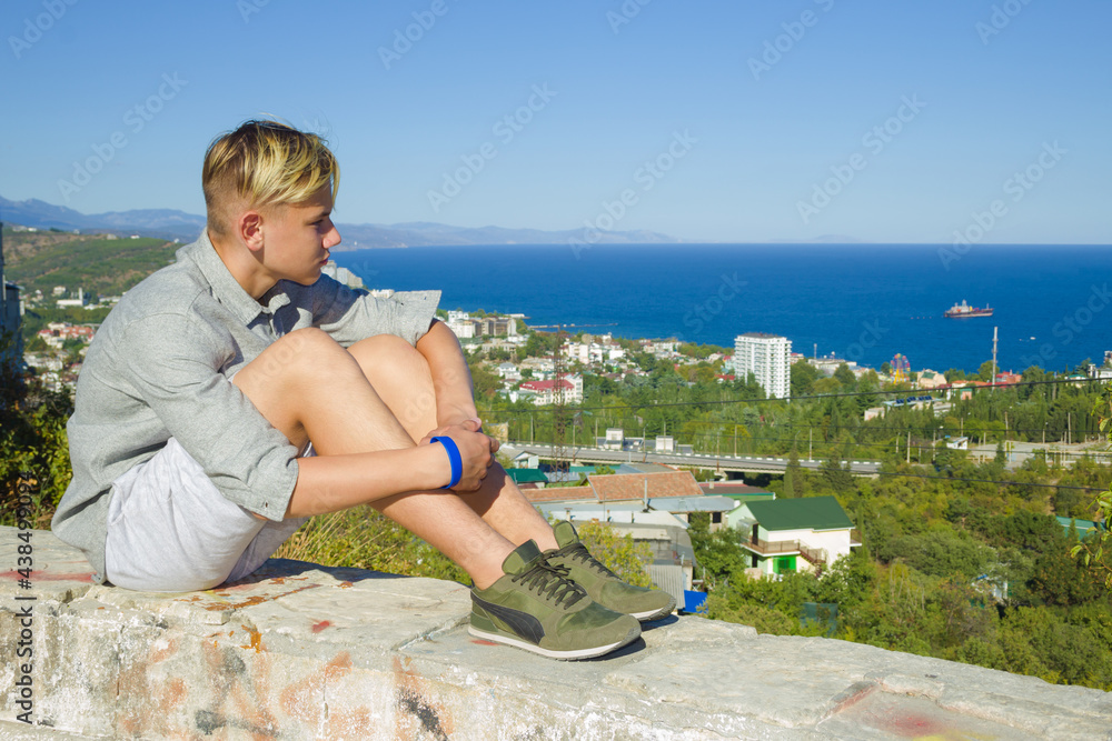 A lonely blond teenager in a gray shirt, shorts, and green sneakers sits on a stone side and looks down at the coastal city of the sea. Blue, cloudless sky and blue sea. A cargo ship leaves the coast