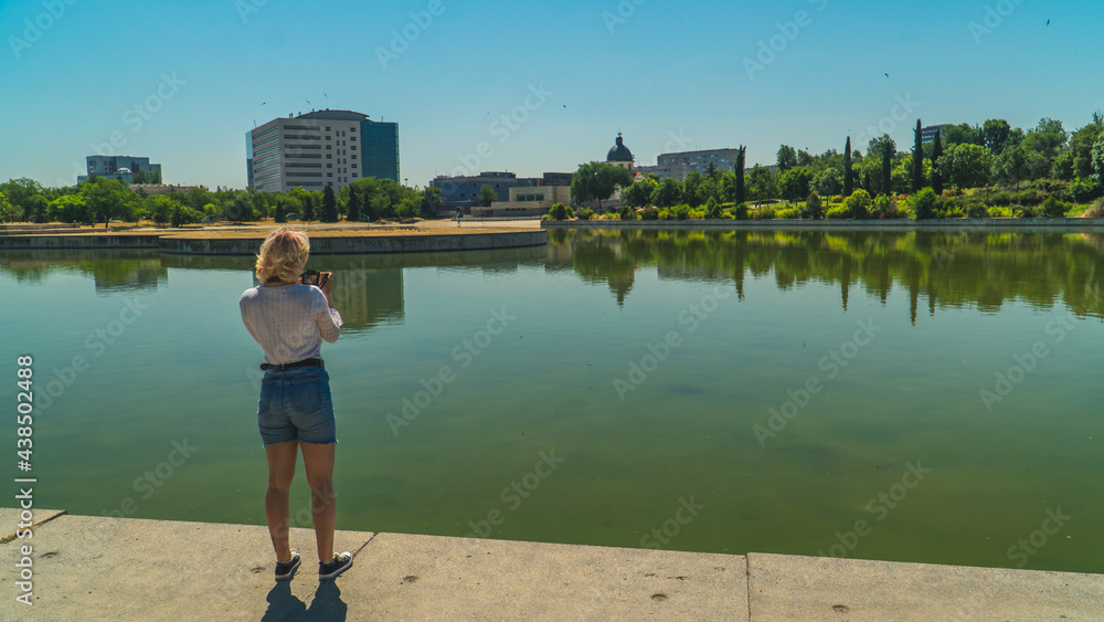 Fototapeta premium mujer mirando fotos en una cámara réflex a lado del lago, mujer rubia con blusa blanca y pantalones cortos azules sujetando una cámara réflex